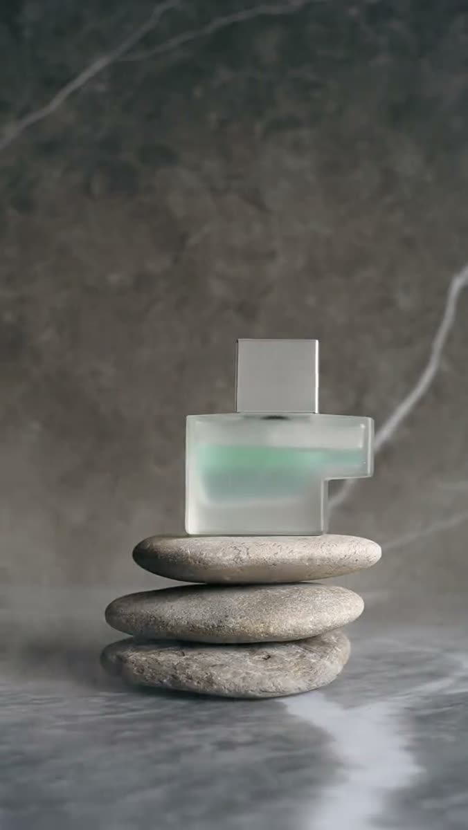 A frosted glass perfume bottle with a mirrored cap balanced on three stacked river stones, against a gray marble wall