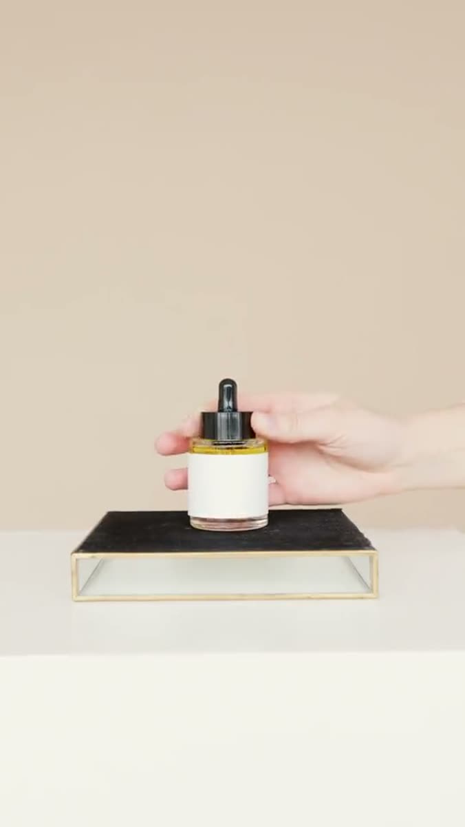 A hand lowering an unlabeled amber dropper bottle onto a black stone tray with a thin brass frame, against a plain beige backdrop