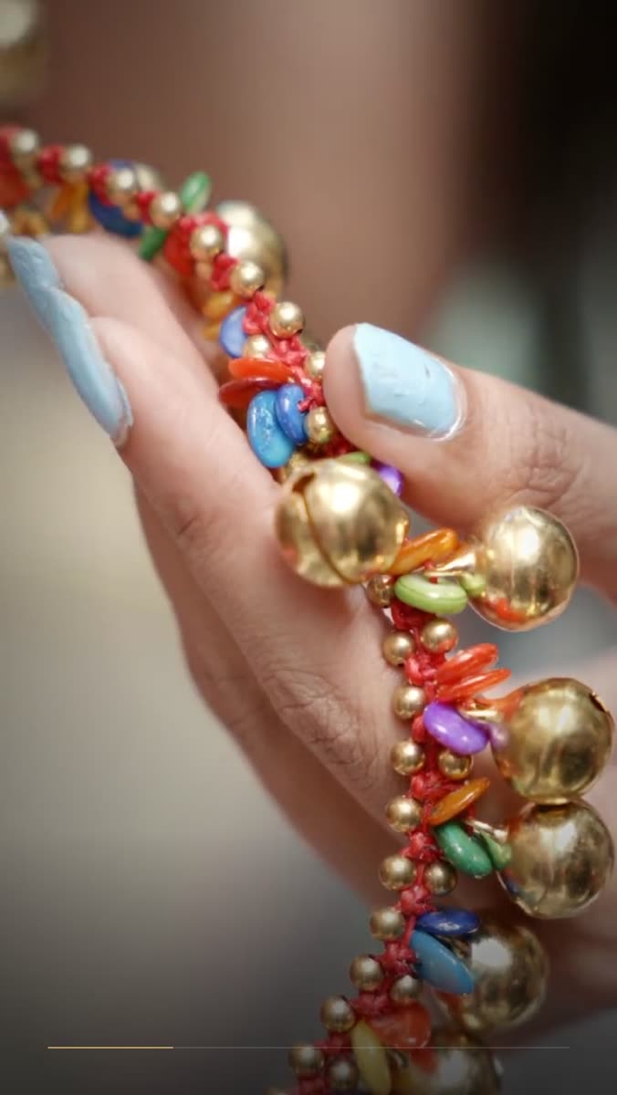 Fingers with pale blue nails holding a strand of gold bells and colored glass beads knotted onto red thread, background thrown out of focus