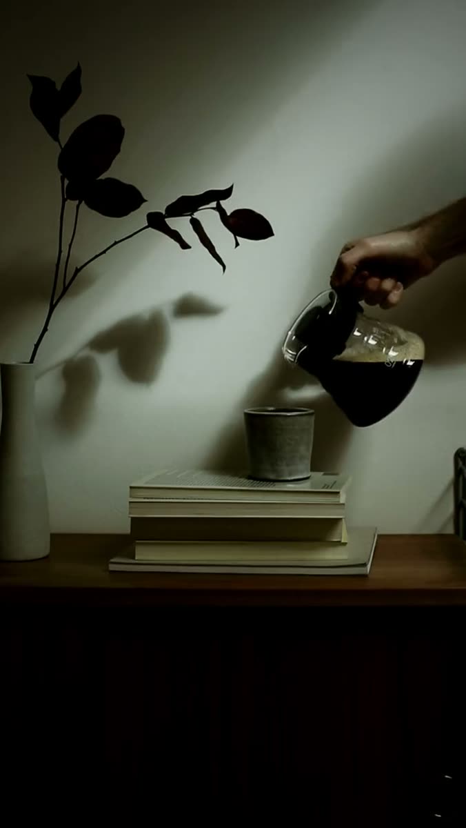 A hand tips a glass carafe of dark coffee toward a stoneware cup balanced on a stack of books, lit by hard low light, with the shadow of a bare branch thrown across the wall behind