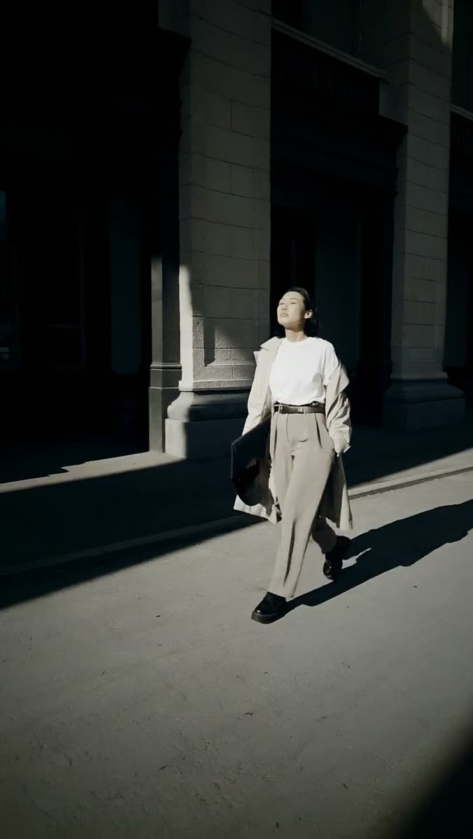 A woman in a white t-shirt and wide beige trousers walking through a hard shaft of sunlight past a dark stone colonnade