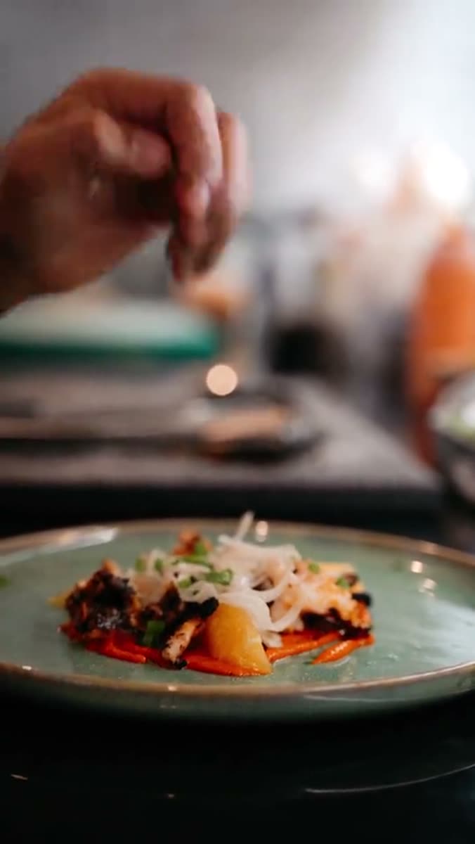 A hand seasoning a plated dish from above, a pale green ceramic plate in the foreground and a blown-out bright window behind, from one of our hotel films